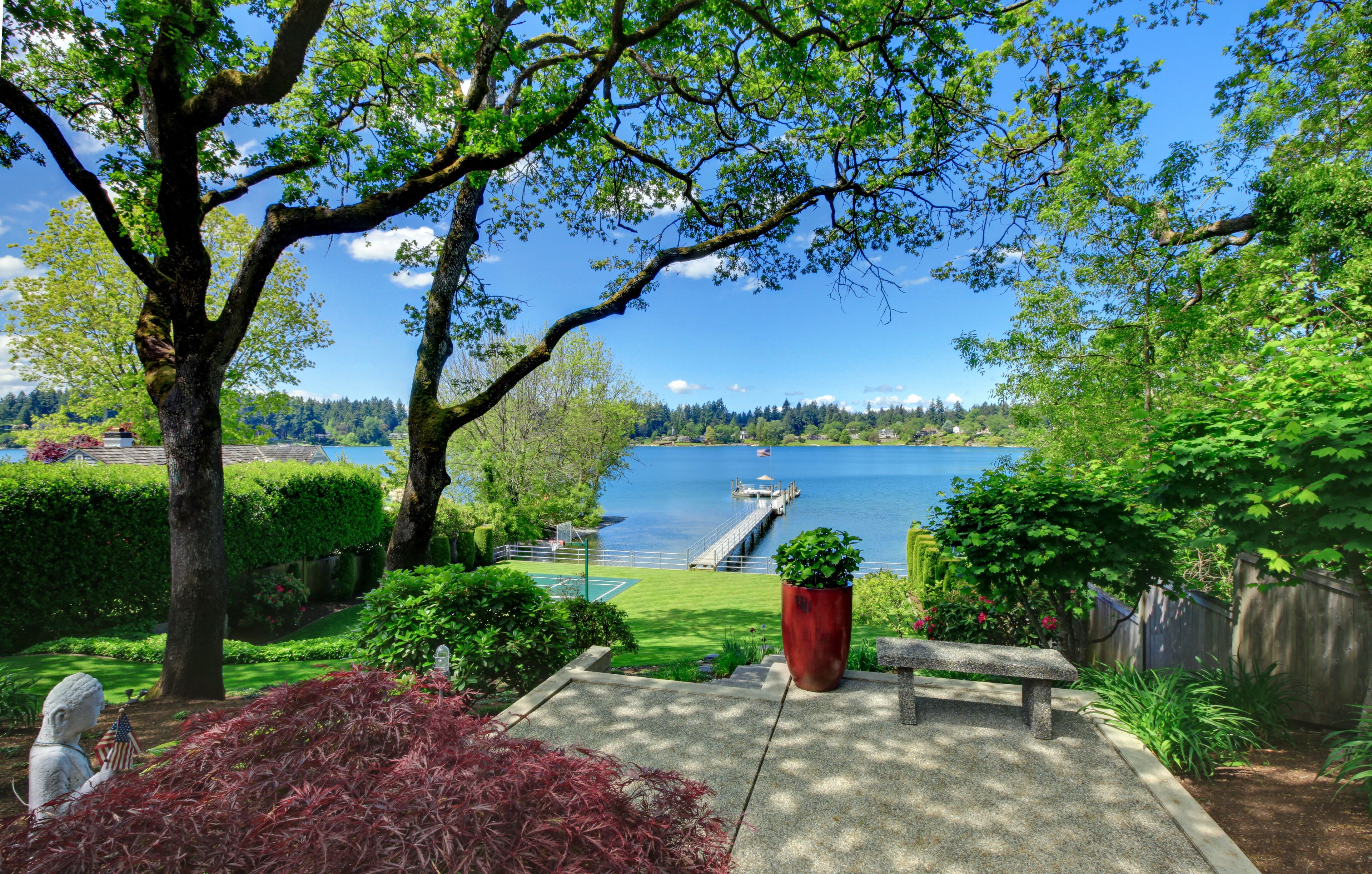A serene lakeside garden with a bench, a large tree, and a view of a dock extending into the water.