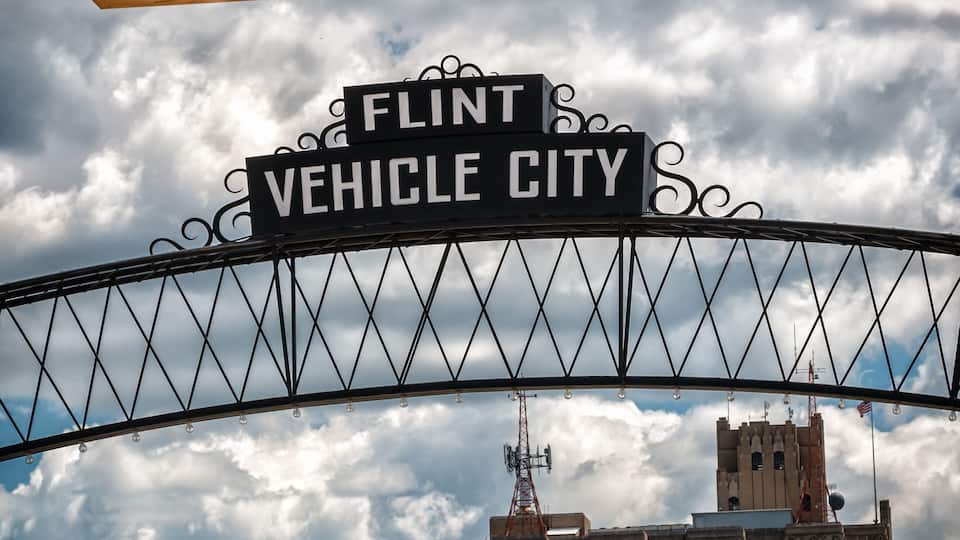 A sign reading 'FLINT VEHICLE CITY' above a bridge with a cloudy sky in the background.