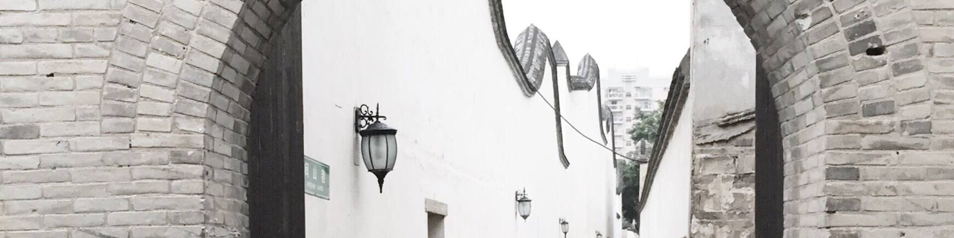 A narrow alleyway with brick walls and white plaster, featuring arched doorways and wall-mounted lanterns.