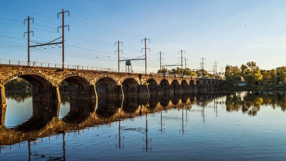 A stone bridge over a calm river with clear reflections, surrounded by trees and power lines.