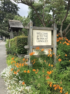 Placa de madeira dando as boas-vindas aos visitantes para o Inn at Langley, rodeada por flores laranjas e brancas vibrantes.