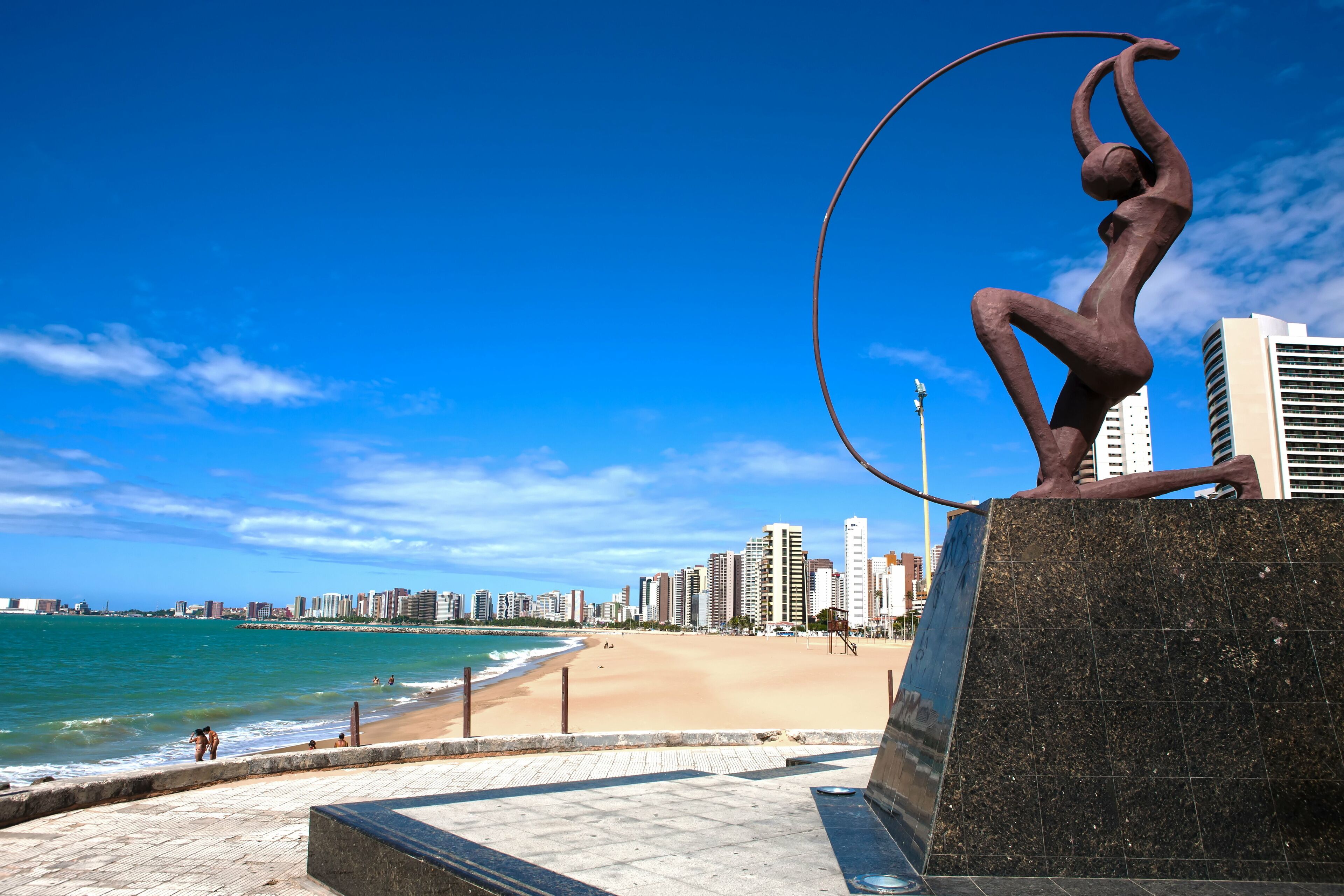 Uma praia com uma escultura, céu azul límpido e um horizonte urbano ao fundo.