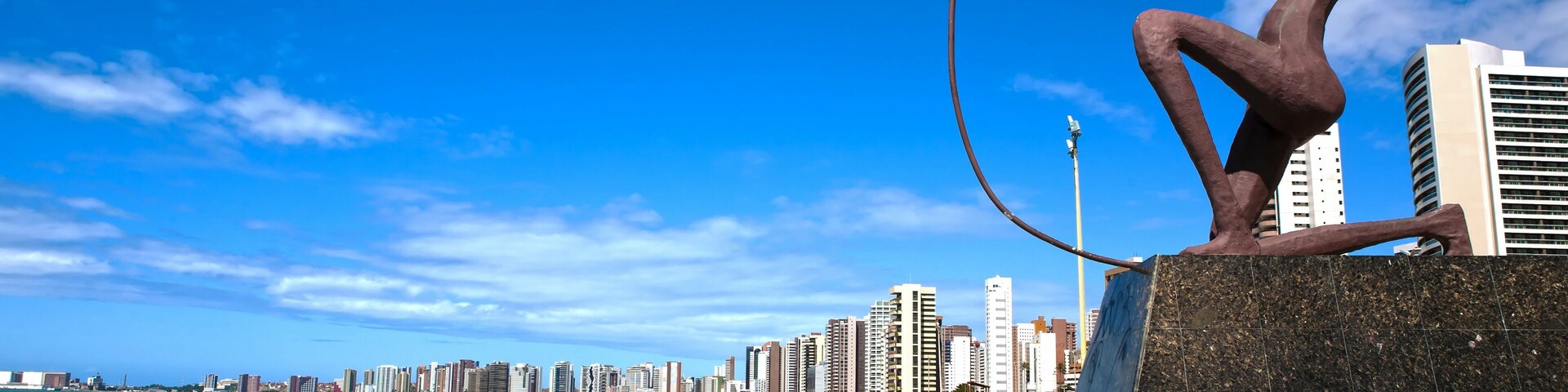 Uma praia com uma escultura, céu azul límpido e um horizonte urbano ao fundo.