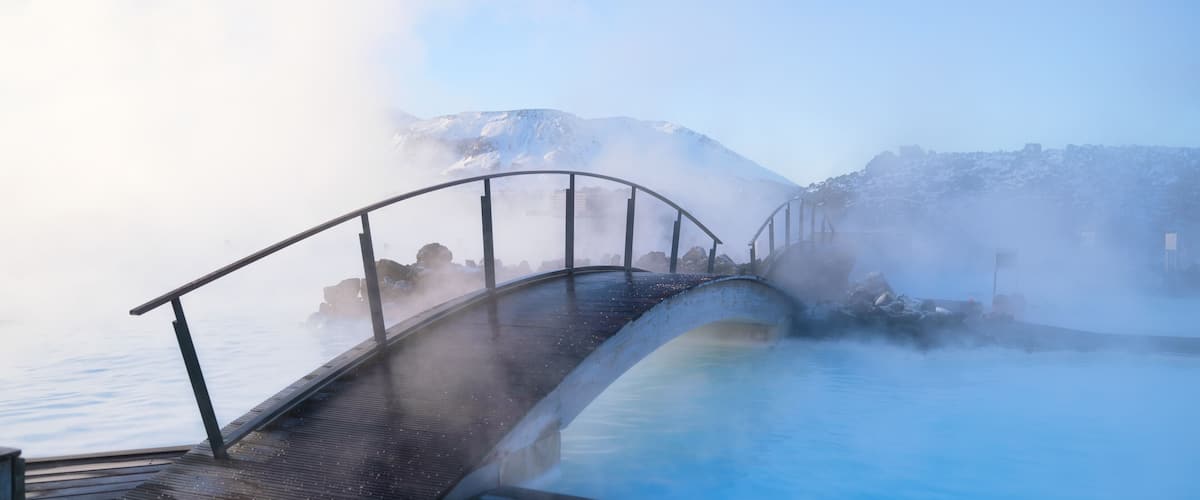 Una terraza de madera con vistas a un manantial termal con vapor ascendente, un puente de metal y montañas nevadas al fondo.