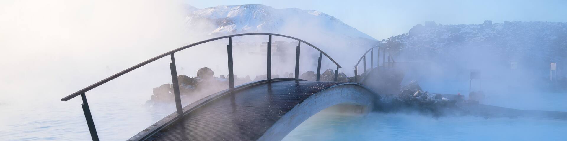 A wooden deck overlooking a hot spring with steam rising, a metal bridge, and snow-capped mountains in the background.