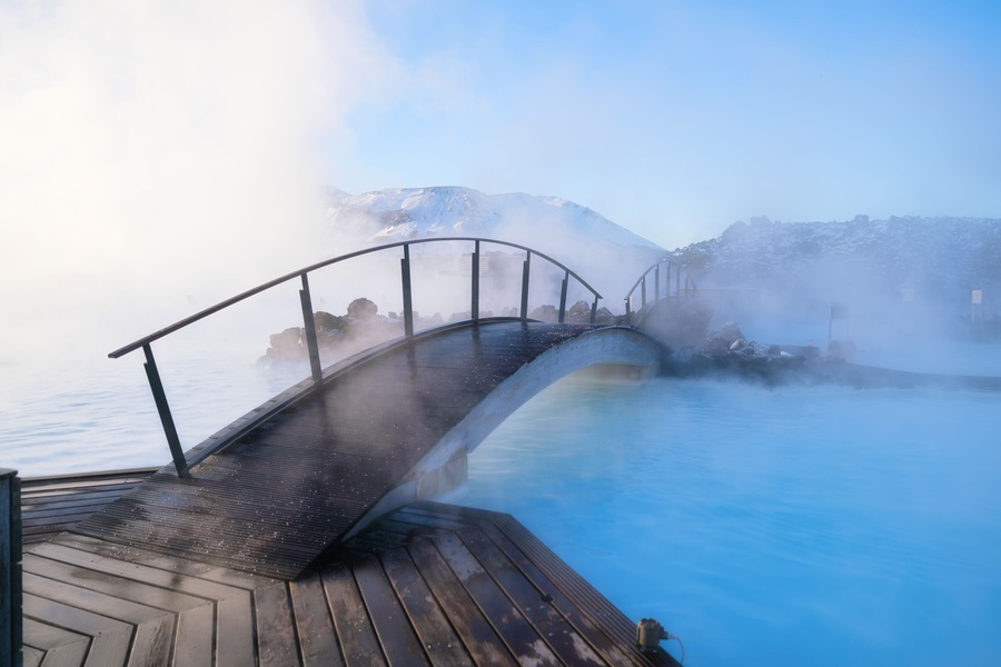Une terrasse en bois surplombant une source thermale où s’élève de la vapeur, un pont métallique et, au loin, des montagnes aux sommets enneigés.