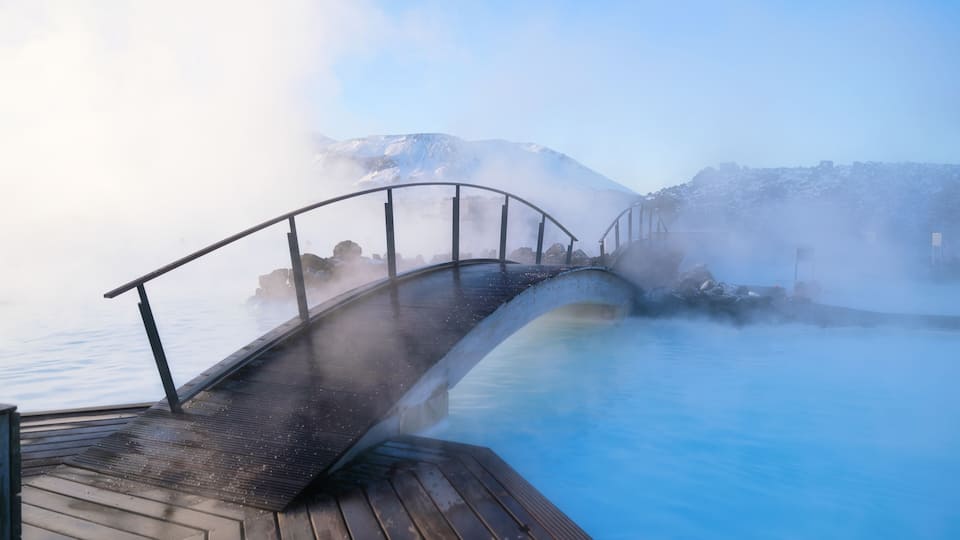 A wooden deck overlooking a hot spring with steam rising, a metal bridge, and snow-capped mountains in the background.