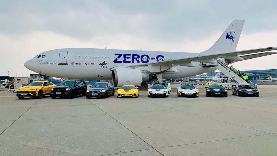 Ein ZERO-G Flugzeug mit den Logos von noveSpace, CSA und DLR auf der Außenhülle, geparkt auf dem Vorfeld eines Flughafens, wobei mehrere Luxusautos davor abgestellt sind.
