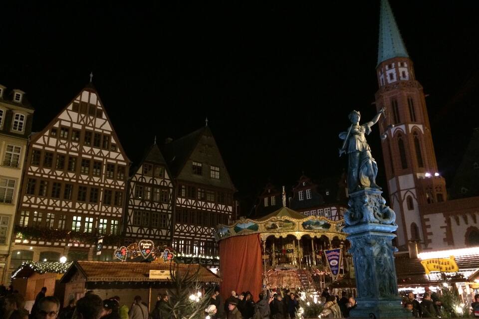 Uma praça tradicional de cidade europeia à noite, com edifícios históricos, uma estátua e um campanário.