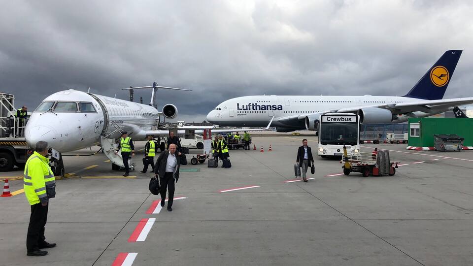 Une aire de stationnement d’aéroport avec des avions de Lufthansa et d’une autre compagnie aérienne, du personnel au sol et un autobus pour l’équipage.