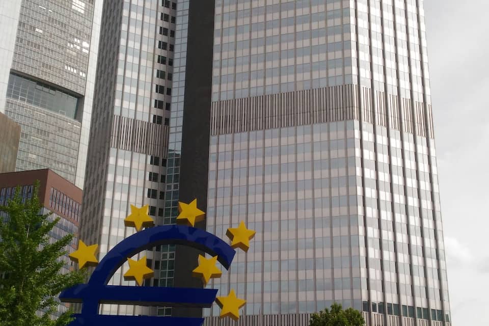Een modern wolkenkrabber met een glazen gevel, een groot sculptuur van de Europese vlag, en mensen lopen op de voorgrond.