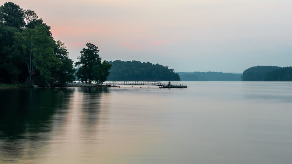 A calm lake with a dock extending into the water, surrounded by dense trees and a distant shoreline.