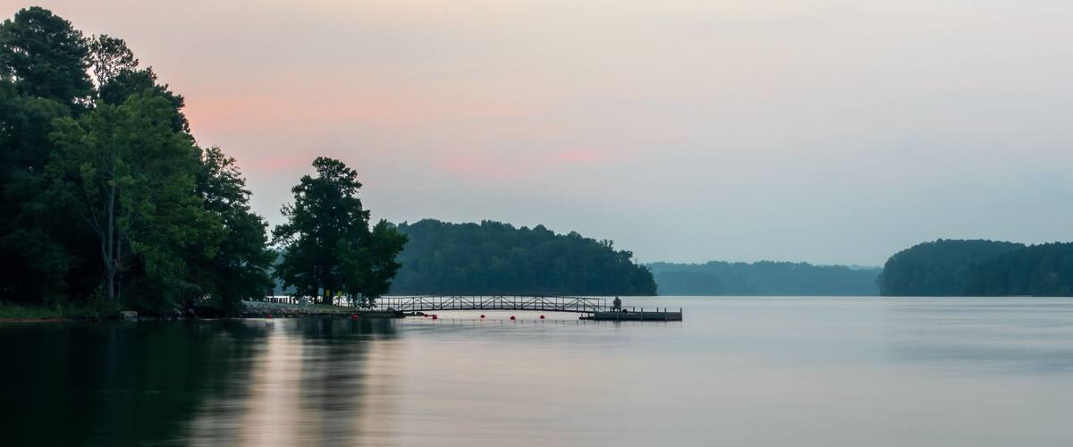 A calm lake with a dock extending into the water, surrounded by dense trees and a distant shoreline.