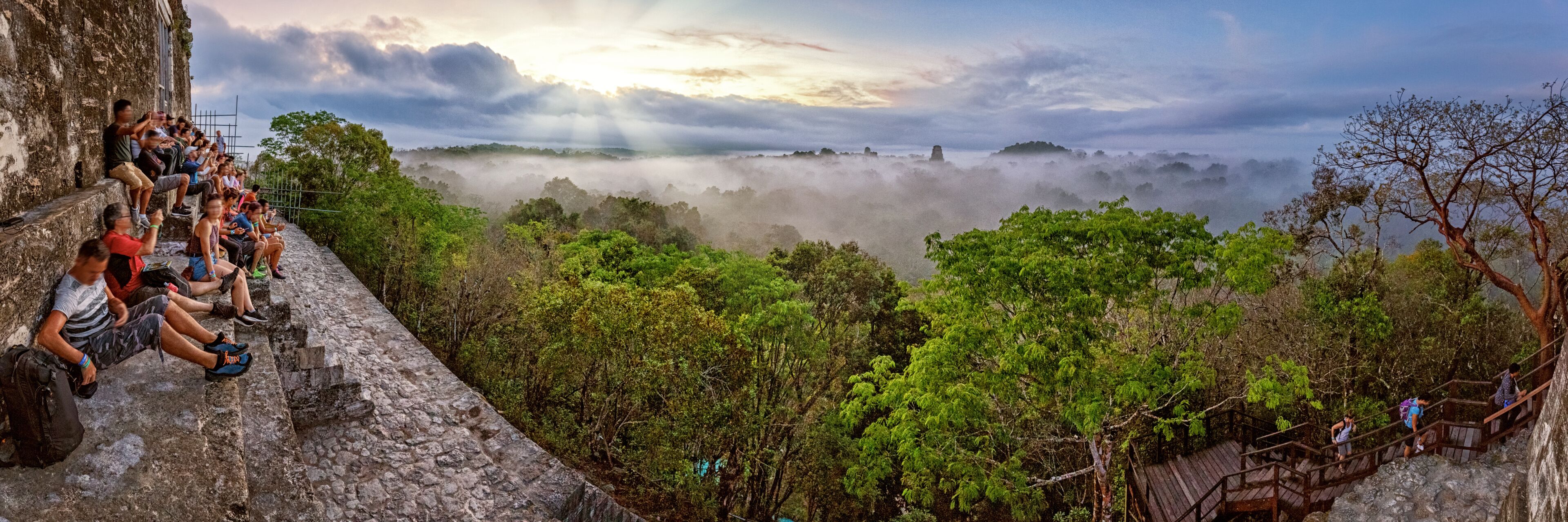 A group of people sitting on a stone ledge overlooking a misty forested valley with a sunrise in the background.