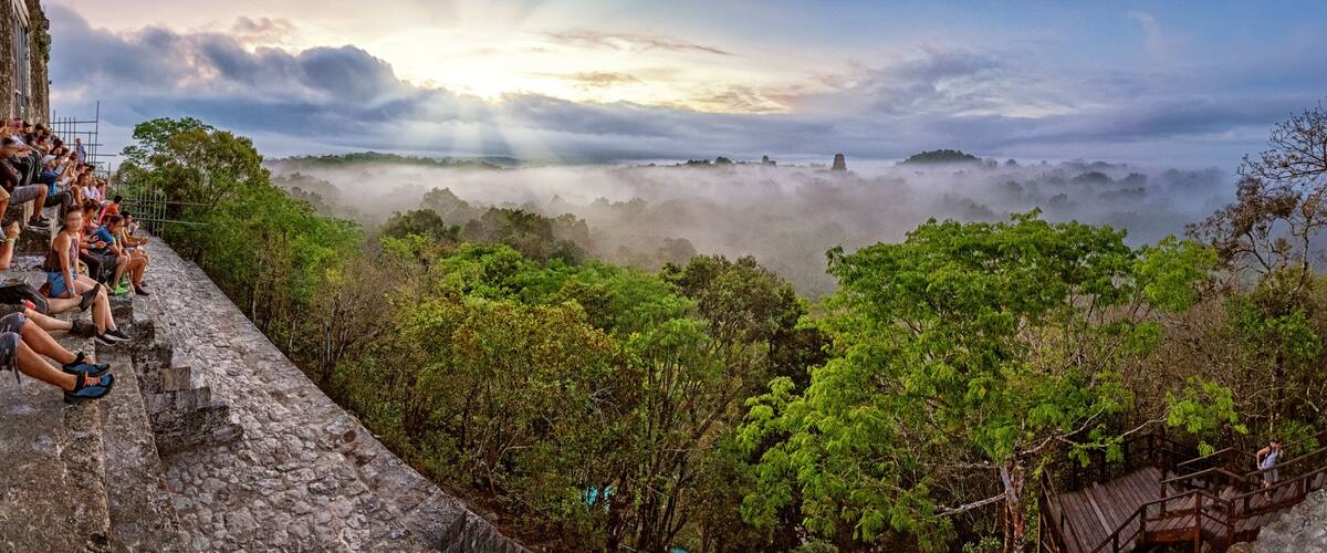 Un gruppo di persone sedute su una sporgenza in pietra con vista su una valle boscosa avvolta dalla nebbia e un'alba sullo sfondo.