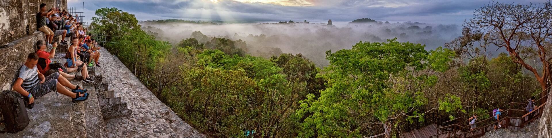 A group of people sitting on a stone ledge overlooking a misty forested valley with a sunrise in the background.