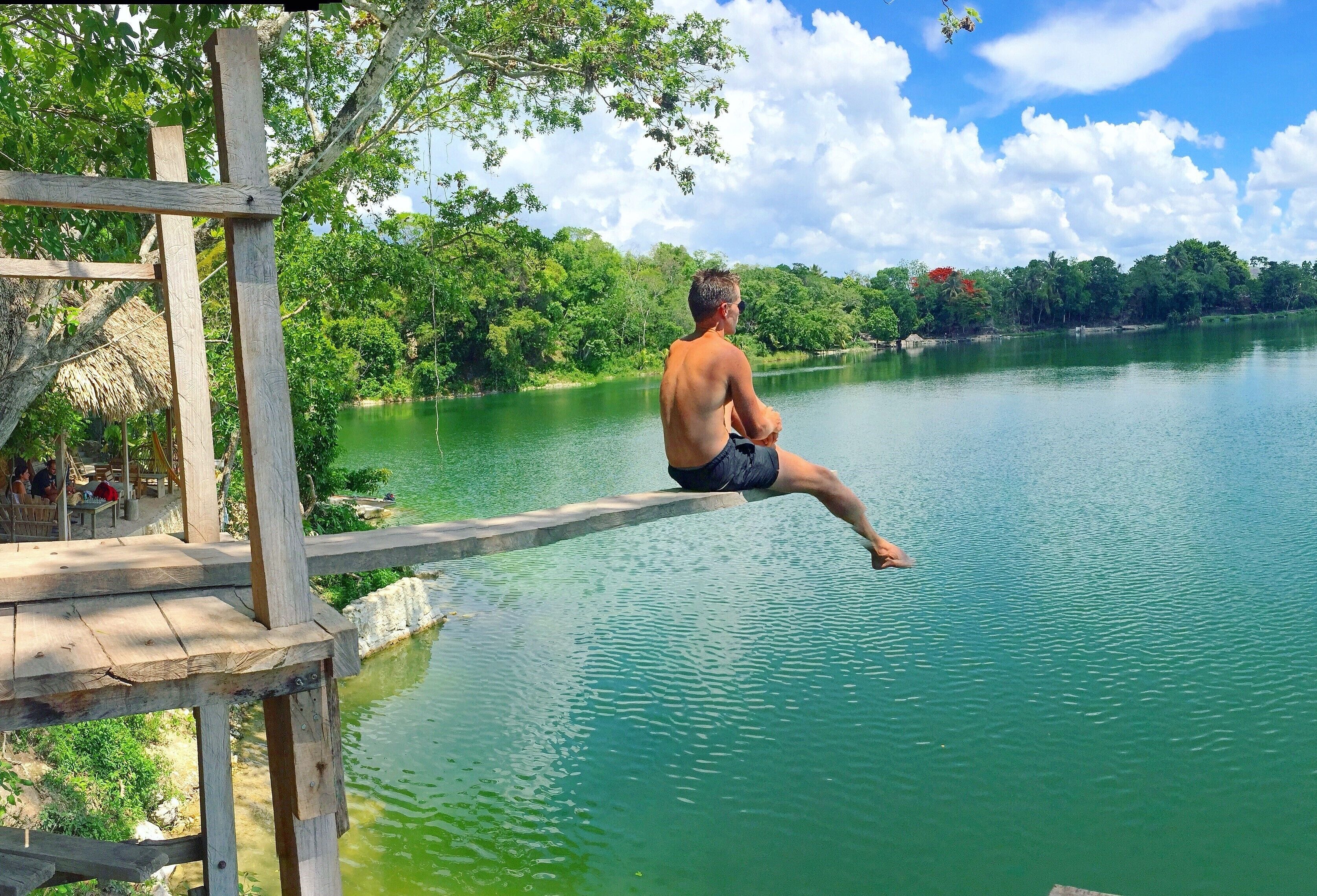 A man sitting on a wooden dock by a calm lake surrounded by lush greenery.
