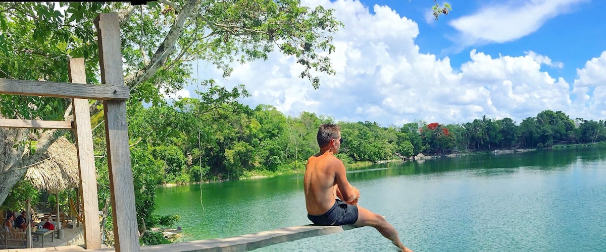 A man sitting on a wooden dock by a calm lake surrounded by lush greenery.
