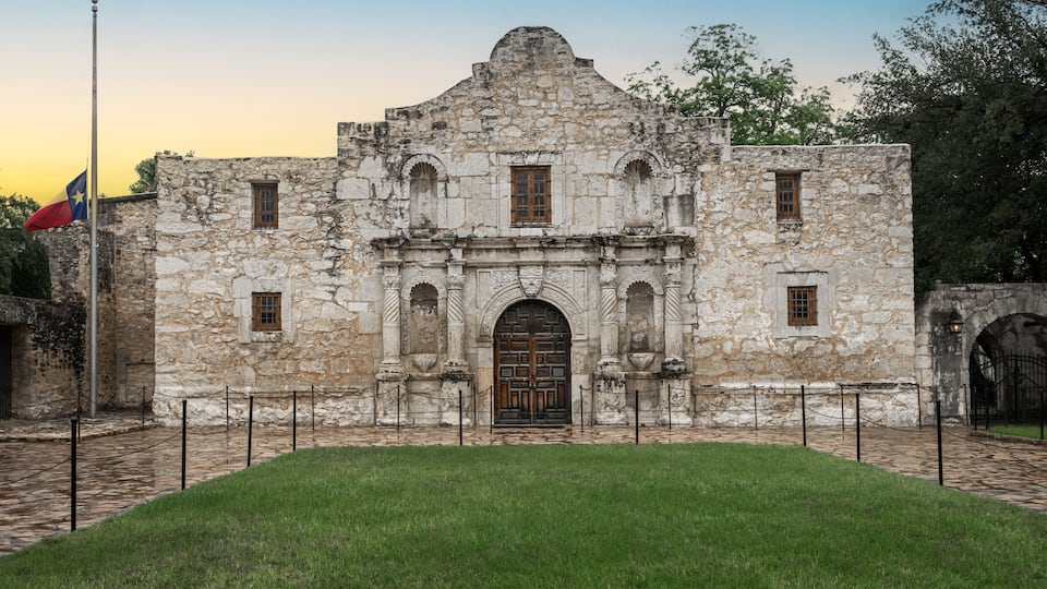 A historic stone building with a central arched doorway, a flagpole with a flag, and a paved courtyard.