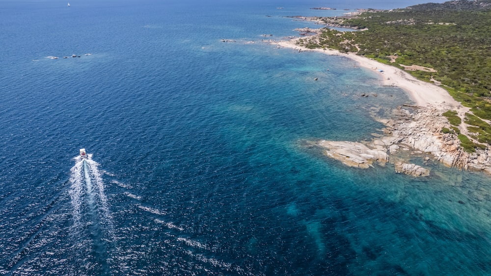 Eine Küstenlandschaft mit klarem, blauem Wasser, einem Sandstrand, Felsformationen und einem entfernten Segelboot.