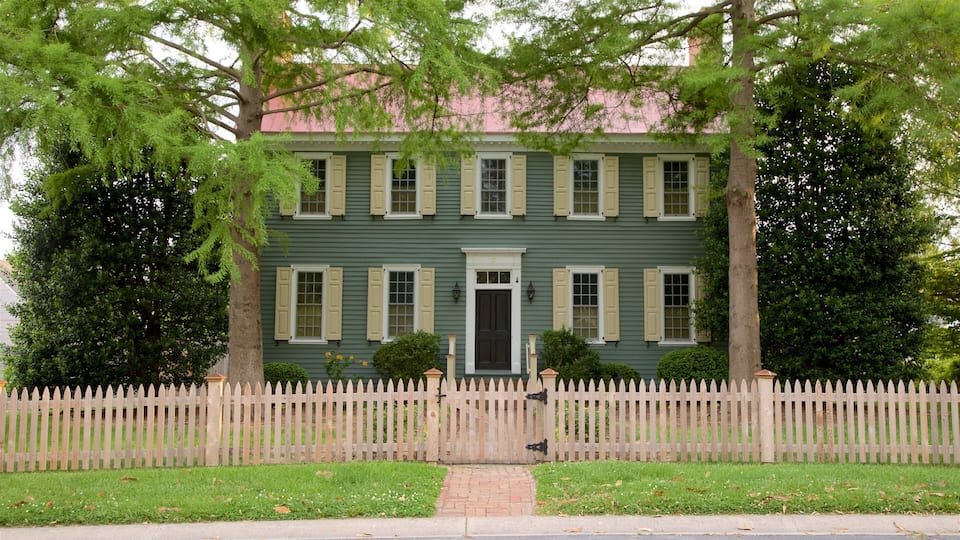 Una casa de dos pisos con fachada verde y contraventanas blancas, rodeada por un cerco blanco de piquetes y árboles.