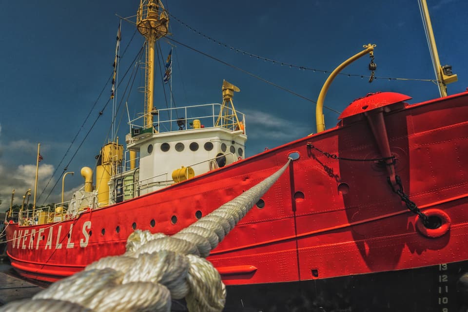Een rode boot genaamd 'Overfalls' ligt aangemeerd in een haven, met een helderblauwe lucht op de achtergrond.