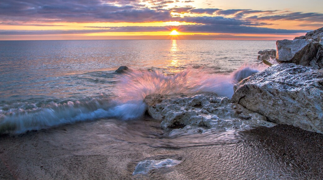 Waves Crashing On Beach. Wave crashing over a rocky coast with sunrise colors reflecting on the waters of Lake Huron In Lexington, Michigan.