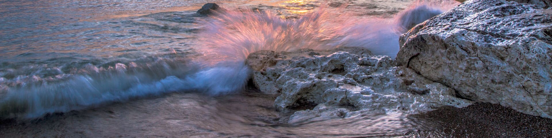 Waves Crashing On Beach. Wave crashing over a rocky coast with sunrise colors reflecting on the waters of Lake Huron In Lexington, Michigan.
