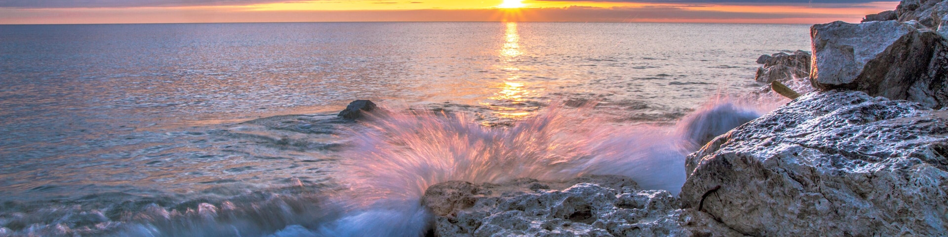 Waves Crashing On Beach. Wave crashing over a rocky coast with sunrise colors reflecting on the waters of Lake Huron In Lexington, Michigan.