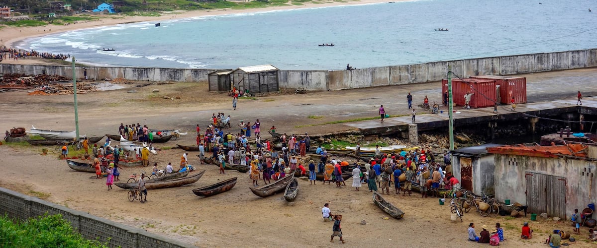 A crowded beach with people, boats, and a mountainous backdrop.