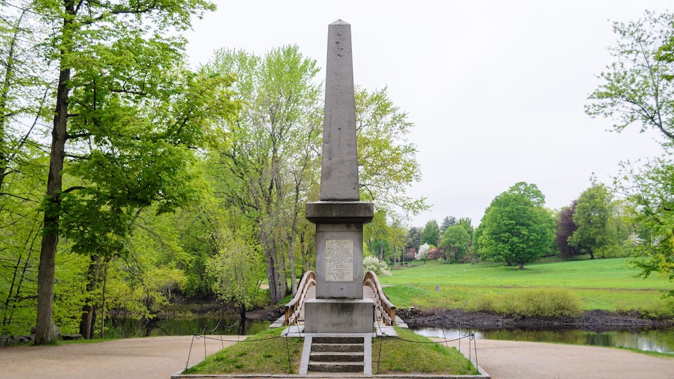 A tall, narrow monument with a plaque in a park with trees and a pond.