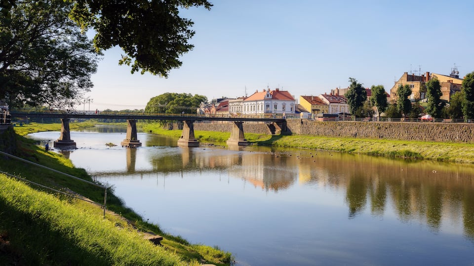 A calm river with a stone bridge, a grassy bank, and buildings in the background.