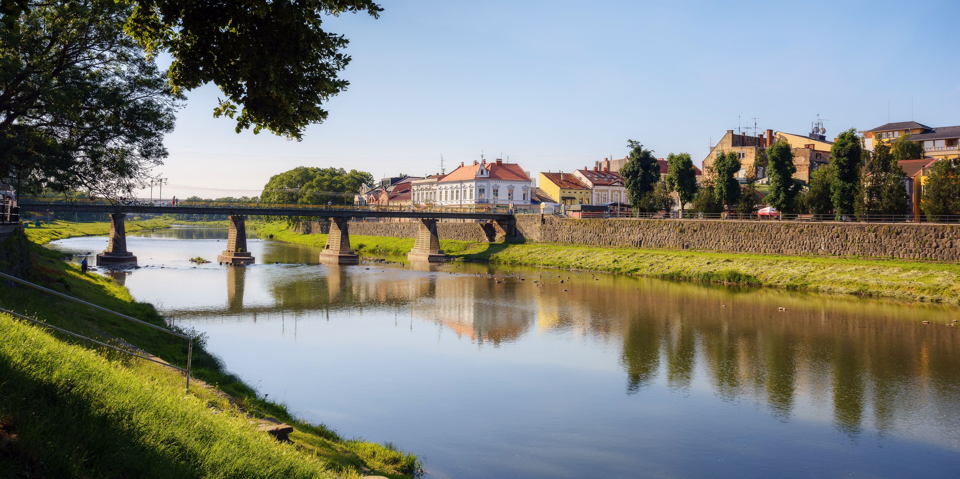 A calm river with a stone bridge, a grassy bank, and buildings in the background.