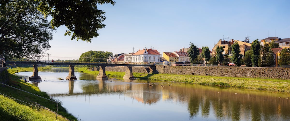 A calm river with a stone bridge, a grassy bank, and buildings in the background.