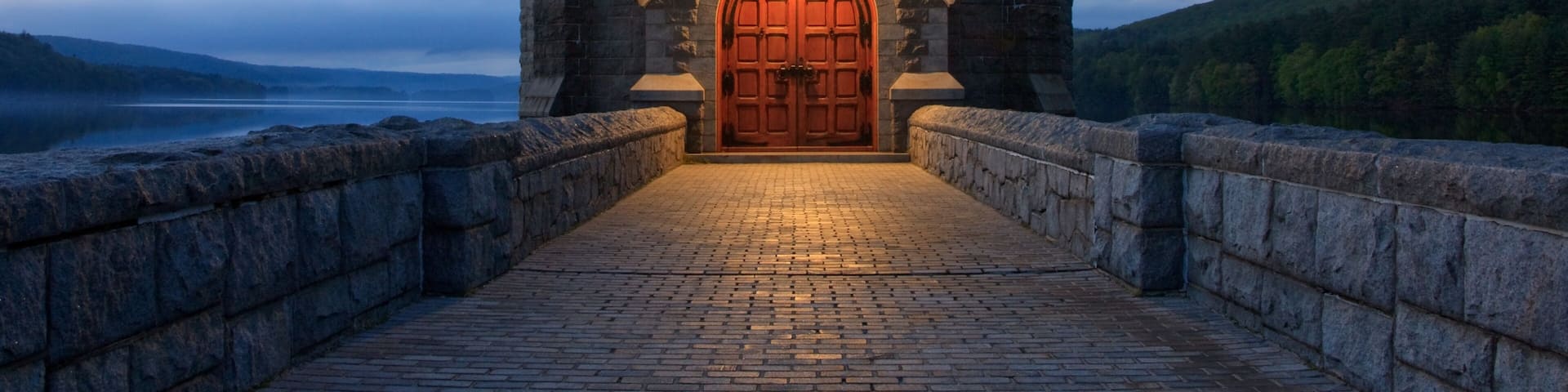 Saville Dam and Reservoir in the evening in Barkhamsted, Connecticut. A walkway leads to a castle tower resting on a lake surrounded by mountains