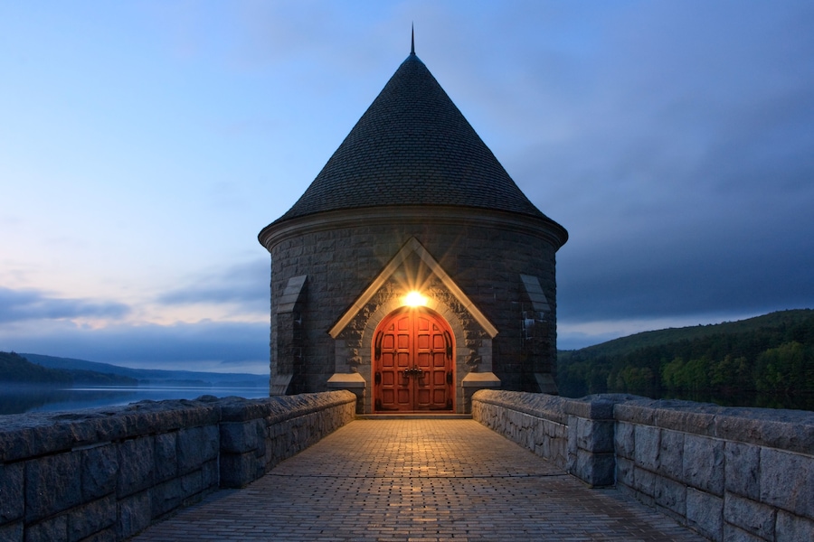 Saville Dam and Reservoir in the evening in Barkhamsted, Connecticut. A walkway leads to a castle tower resting on a lake surrounded by mountains