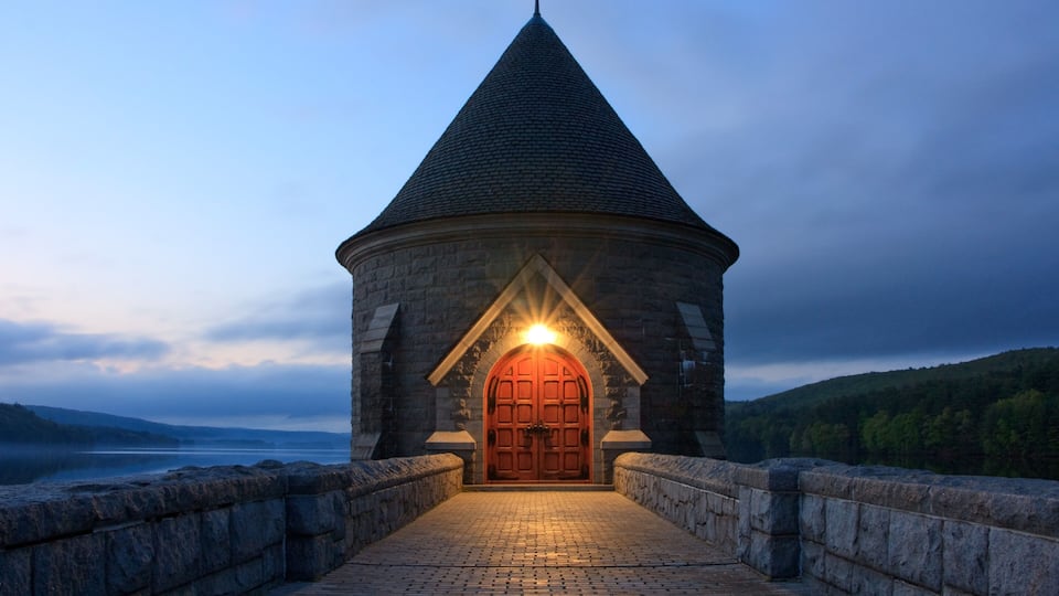 A stone tower with a conical roof and a red door, surrounded by a stone wall and a cobblestone path, overlooking a body of water and hills.