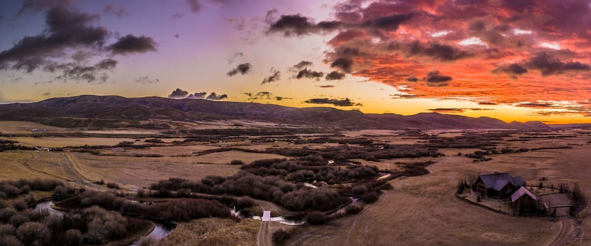 A rural landscape with a house, rolling hills, a river, and a sunset.