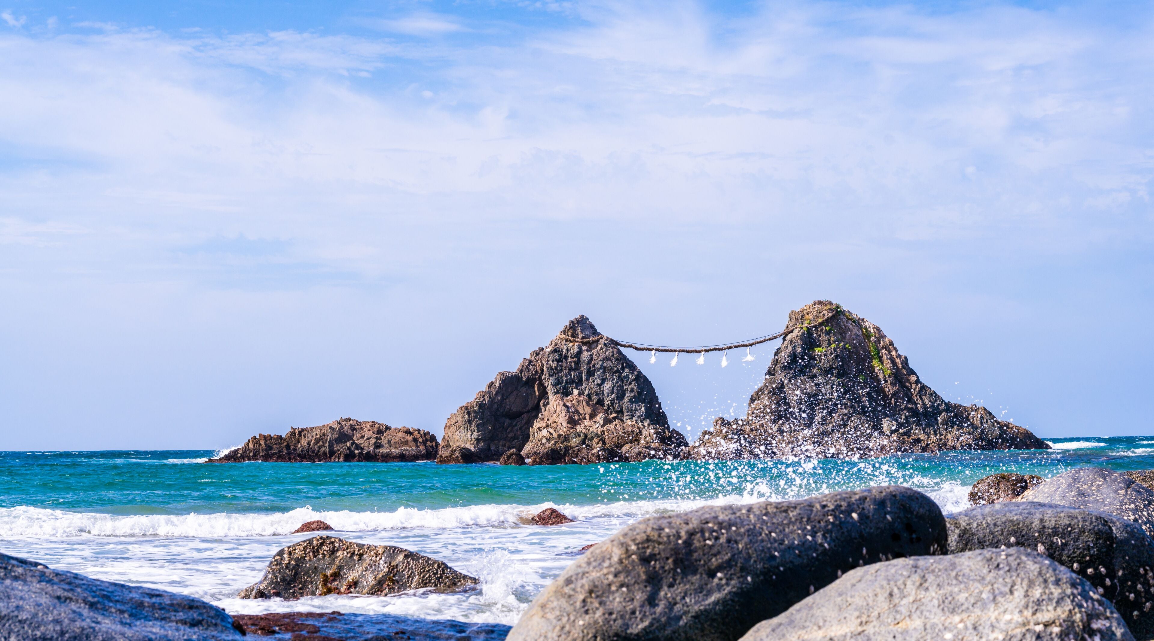 Una costa rocosa con dos formaciones rocosas destacadas unidas por un puente colgante sobre aguas turquesas.