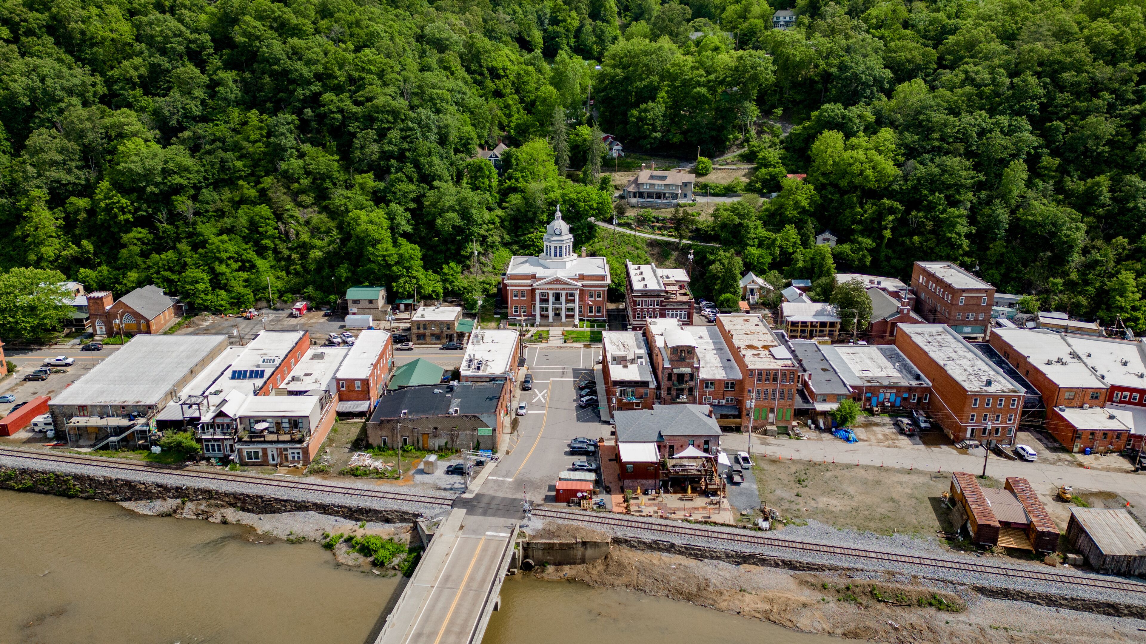 An aerial view of a town with a prominent red-brick courthouse, a railway crossing, and dense forest in the background.