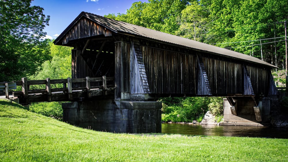 Weathered wooden country covered bridge on a bright sunny day, trees and grass surrounding the bridge. Blue sky with a single cloud above. Livingston Manor, New York State.