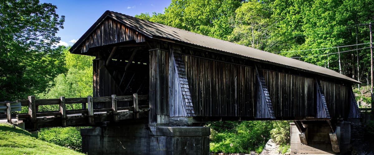 Weathered wooden country covered bridge  on a bright sunny day, trees and grass surrounding the bridge. Blue sky with a single cloud above. Livingston Manor, New York State.