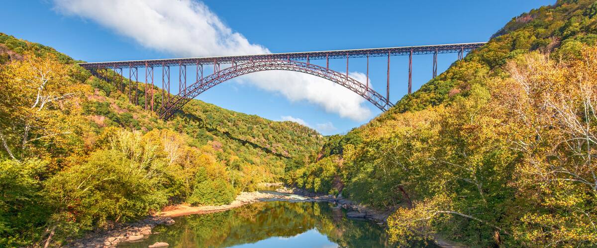 A steel bridge spanning a river, surrounded by autumn foliage and hills.