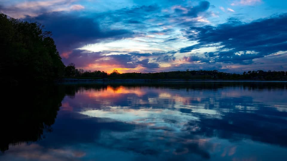 A sunset over a calm lake with a silhouette of trees and a cloudy sky.