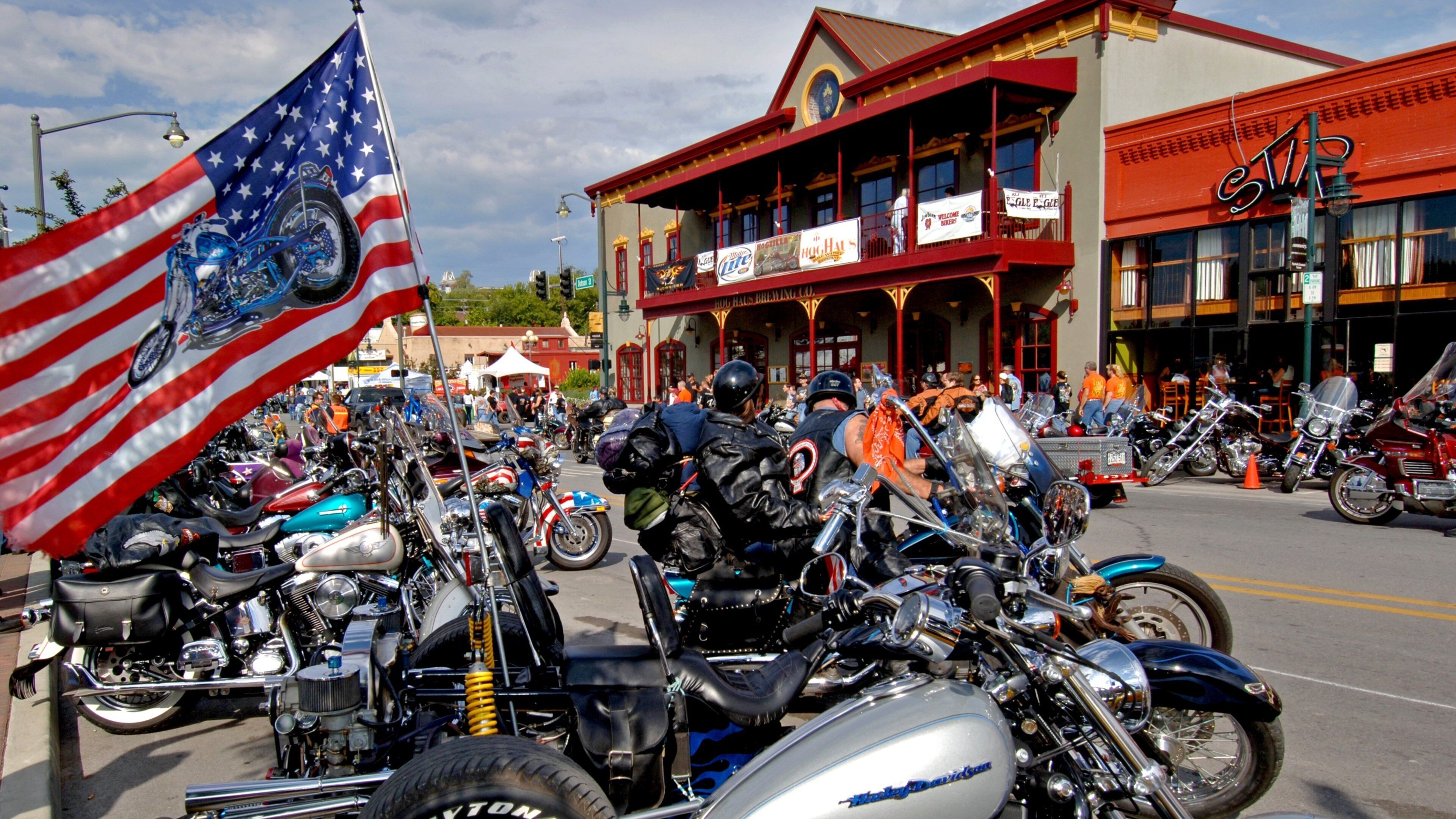 A motorcycle rally with a large American flag, vintage cars, and a historic building with banners.