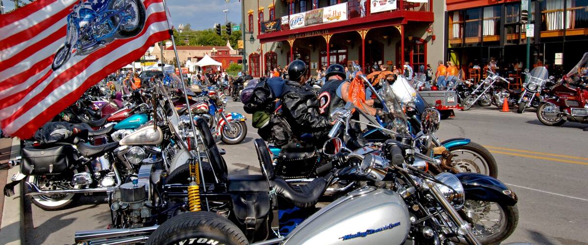 A motorcycle rally with a large American flag, vintage cars, and a historic building with banners.