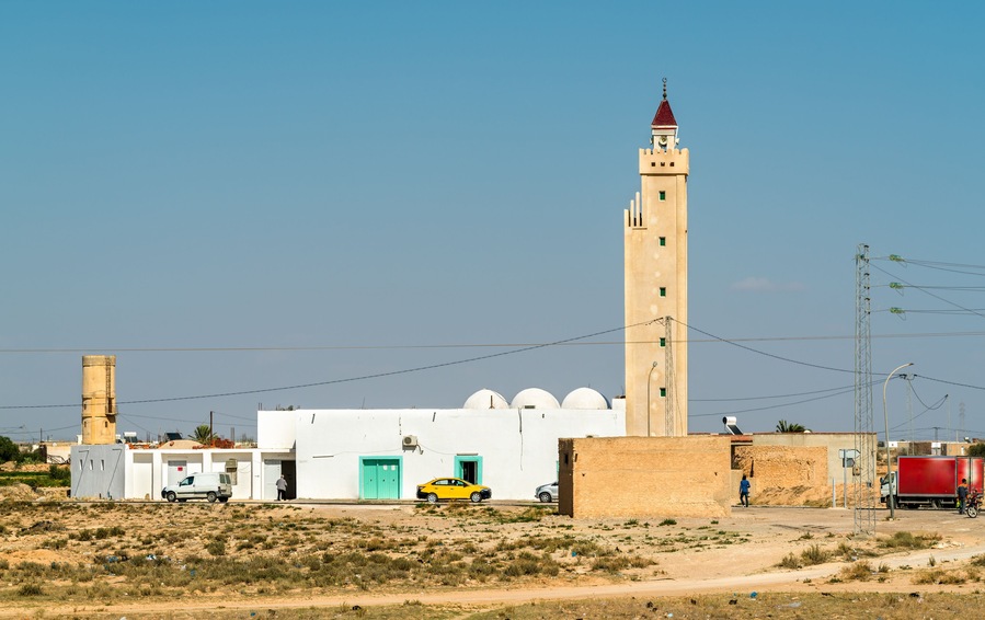 Una mezquita con un minarete alto, un edificio blanco con una puerta verde, y un coche amarillo estacionado en la entrada.