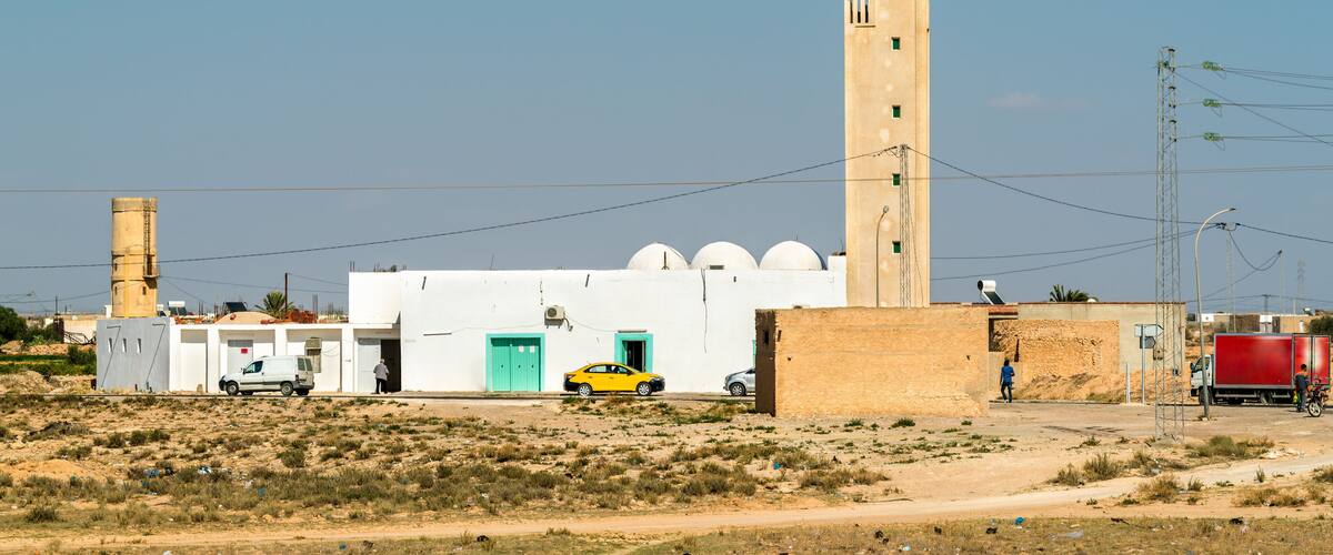 A mosque with a tall minaret, a white building with a green door, and a yellow car parked in front.