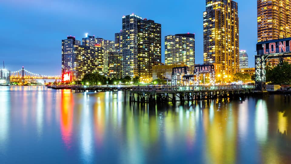 A city skyline at dusk with illuminated buildings reflecting on the water, a bridge, and a sign reading 'LONG ISLAND.'