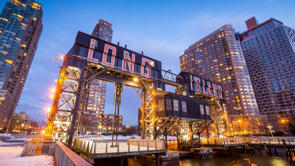 A steel bridge with 'LONG ISLAND' written on it, surrounded by tall buildings and a body of water.